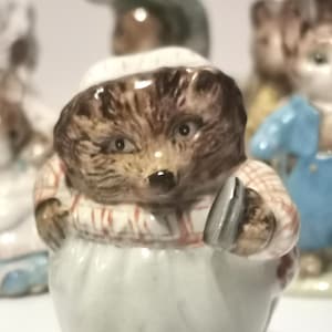 Puede incluir: Figura de cerámica de un erizo con gorro y delantal de chef blancos, sosteniendo un utensilio plateado. El erizo tiene pelaje marrón y viste una camisa a cuadros rojos y blancos. Otras figuras son visibles en el fondo.