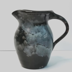 May include: A ceramic jug with a dark, glossy finish. The jug has a rounded body, a curved handle, and a spout for pouring. The color transitions from black to a cloudy blue-grey, creating a unique, artistic look. Suitable for serving beverages.