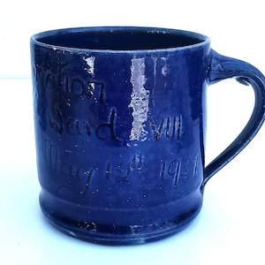 May include: A dark blue ceramic mug with a handle. The mug features etched text: "Ward VIII May 12th 1931". The mug has a glossy finish and a slightly worn look. A vintage item.