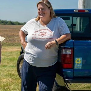 May include: A light gray t-shirt with the text "The Wiener Hole" in red and white, featuring a hot dog graphic. The shirt also includes the phrase "Grab a frank with a friend!" and the words "Established 1979". The person is wearing navy blue pants.