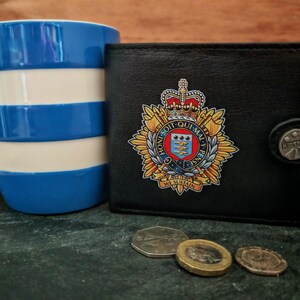 May include: A black leather wallet with a crest emblem, a blue and white striped mug, and several coins are displayed. The wallet has a snap closure and the crest is in gold, red, and blue. The mug is ceramic.