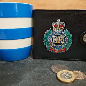 May include: A black leather wallet featuring the Royal Engineers emblem with the text Honi Soit Qui Mal Y Pense and ER, alongside a blue and white striped mug and three coins. The wallet has a snap closure with the brand name Klassek visible.