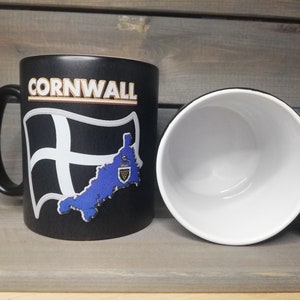 May include: Black ceramic mug with a white and blue design featuring the text "CORNWALL" and a stylised map of Cornwall with a flag. The mug has a white interior.