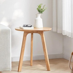 May include: A round, light-colored wooden side table with three angled legs. A white vase with a textured design holds greenery. A book and eyeglasses rest on the table's surface. The table is approximately 24 inches in diameter.