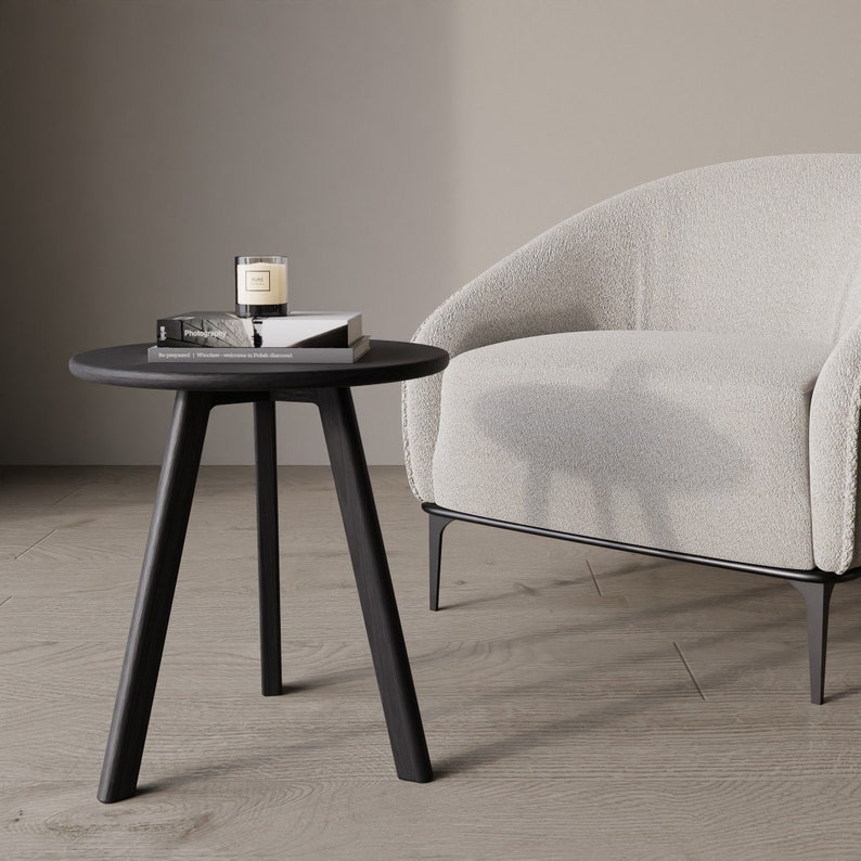 Puede incluir: Una mesa auxiliar negra con tres patas y una tapa redonda. Un libro y una vela est&aacute;n encima de la mesa. Un sill&oacute;n blanco con un marco de metal negro est&aacute; en el fondo.