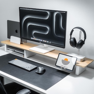 May include: A modern desk setup featuring a large computer monitor with a black and white abstract design. A wooden monitor stand holds a laptop, tablet, and headphones. A keyboard and mouse sit on a black desk mat.