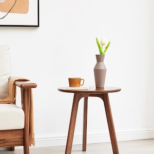 May include: A round wooden side table with a brown finish and three legs. A white tulip in a gray vase and a brown ceramic mug are on the table.