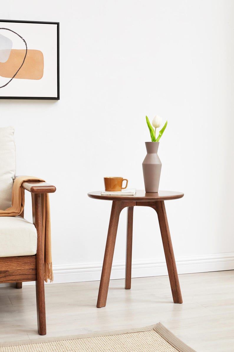 May include: A round wooden side table with a brown finish and three legs. A white tulip in a gray vase and a brown ceramic mug are on the table.