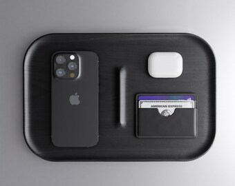 Könnte beinhalten: Ein schwarzes Tablett mit einem schwarzen iPhone, einem weißen kabellosen Ladecase und einem schwarzen Lederkartenhalter mit einer American Express Karte.