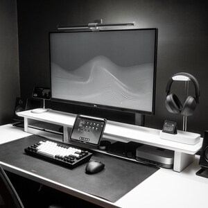 May include: A white desk with a black keyboard, mouse, and headphones. A monitor with a white and grey abstract design is on a white stand. A tablet is on the stand next to the monitor. There is a black speaker on the right side of the desk.
