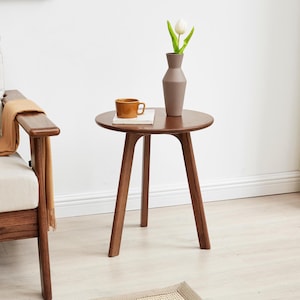 Puede incluir: Una mesa auxiliar redonda de madera con tres patas. La mesa es de color marr&oacute;n y tiene un acabado liso. Un jarr&oacute;n marr&oacute;n con una tulipa blanca y una taza est&aacute;n sobre la mesa.