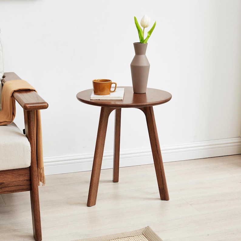 Puede incluir: Una mesa auxiliar redonda de madera con tres patas. La mesa es de color marr&oacute;n y tiene un acabado liso. Un jarr&oacute;n marr&oacute;n con una tulipa blanca y una taza est&aacute;n sobre la mesa.
