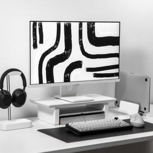 May include: A white desk with a black and white abstract design on the computer monitor. A black headset is on a stand to the left of the monitor. A white keyboard and a white mouse are on the desk.