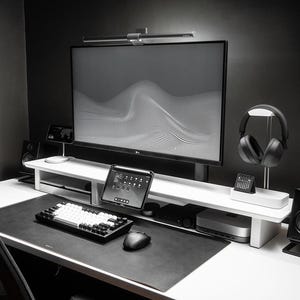 May include: A modern, minimalist workspace featuring a large computer monitor with a wave pattern, a white keyboard, mouse, tablet, and headphones. The setup includes a desk lamp and speakers, all in shades of black and white.