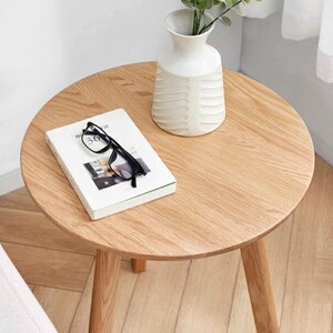 Puede incluir: Una mesa auxiliar redonda de madera con un acabado de madera clara. Un jarr&oacute;n de cer&aacute;mica blanco con un dise&ntilde;o texturizado se encuentra sobre la mesa, lleno de tallos de plantas verdes. Un libro y un par de gafas negras tambi&eacute;n est&aacute;n sobre la mesa.