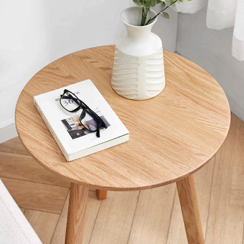 Puede incluir: Una mesa auxiliar redonda de madera con un acabado de madera clara. Un jarr&oacute;n de cer&aacute;mica blanco con un dise&ntilde;o texturizado se encuentra sobre la mesa, lleno de tallos de plantas verdes. Un libro y un par de gafas negras tambi&eacute;n est&aacute;n sobre la mesa.