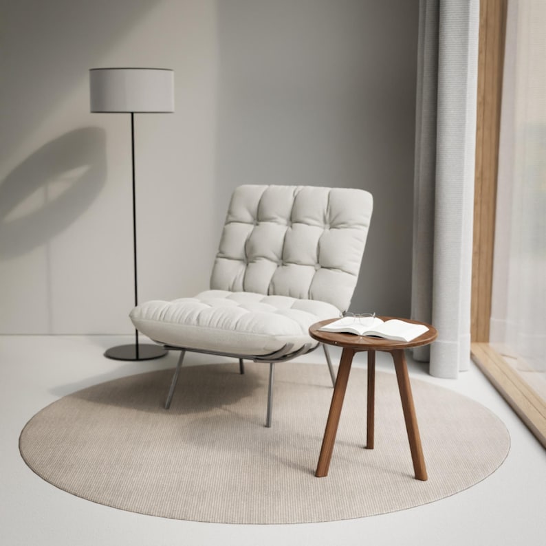 Puede incluir: Un sill&oacute;n tapizado blanco con respaldo capiton&eacute; y una alfombra redonda de color marr&oacute;n claro debajo. Una peque&ntilde;a mesa auxiliar de madera con un libro encima se encuentra junto al sill&oacute;n. Una l&aacute;mpara de pie con una pantalla blanca se encuentra en el fondo.