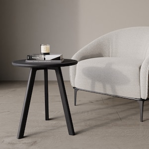May include: A black side table with three legs and a round top. A book and a candle are on the table. A white armchair with a fluffy texture is in the background.