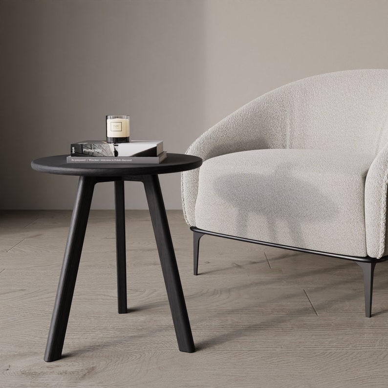 May include: A black side table with three legs and a round top. A book and a candle are on the table. A white armchair with a fluffy texture is in the background.