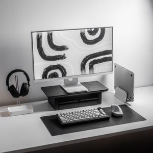 May include: A modern desk setup featuring a computer monitor displaying abstract black brushstrokes on a white background. Includes a black keyboard, mouse, and a black desk mat. A pair of black headphones rests on a white stand.