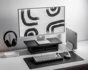 Könnte beinhalten: Moderne Schreibtischeinrichtung mit einem Computermonitor, der abstrakte schwarze Pinselstriche auf weißem Hintergrund zeigt. Enthält eine schwarze Tastatur, Maus und eine schwarze Schreibtischunterlage. Ein schwarzer Kopfhörer liegt auf einem weißen Ständer.