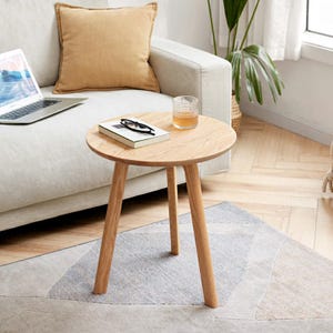 May include: A light wood side table with a round top and three legs. The table is sitting on a gray rug with a geometric pattern.