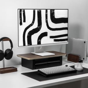 May include: A modern desk setup featuring a white monitor displaying a black and white abstract design. A keyboard, mouse, and laptop are on the desk. Black headphones rest on a wooden stand, and a wooden laptop stand is also present.