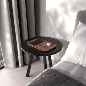 May include: A wooden charging tray with a black phone, white wireless earbuds, and two rings on a black side table next to a bed.