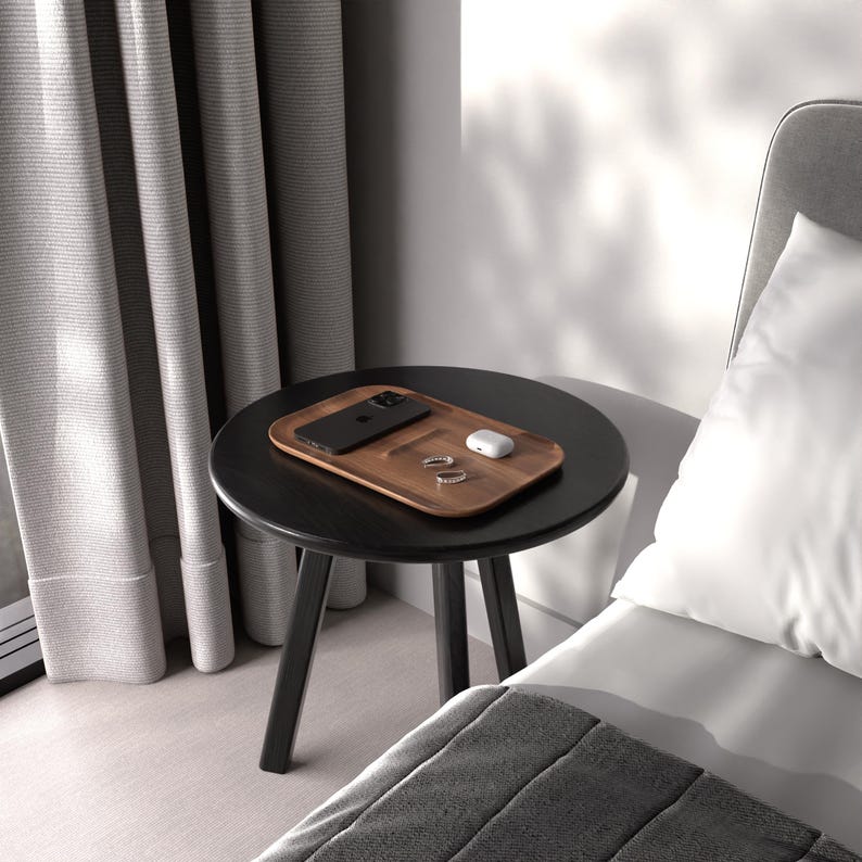 May include: A wooden charging tray with a black phone, white wireless earbuds, and two rings on a black side table next to a bed.