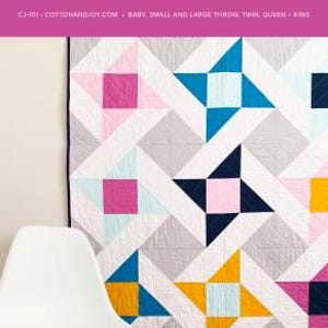 May include: A colorful quilt with a geometric star pattern in shades of pink, blue, yellow, and navy on a gray background. The text "MIGHTY STARS" and "COTTON + JOY" are at the top. A white chair is in the foreground.