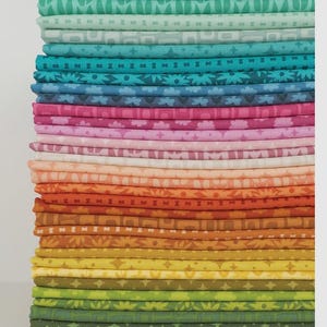 May include: A vertical stack of folded fabric squares arranged in a gradient color spectrum. The textiles feature various repeating patterns including geometric shapes, stars, and floral motifs in shades of green, blue, pink, orange, and yellow.