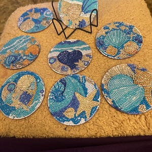 May include: A set of seven round coasters with a beach theme. Each coaster features a unique design with seashells, starfish, and ocean waves in shades of blue, beige, and gold. Includes a black metal holder.