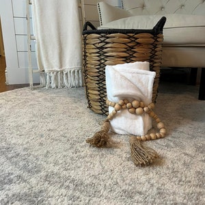 May include: A woven basket with a black rim and handles, filled with white towels and a wooden bead garland with tassels. The basket is sitting on a grey and white patterned rug.
