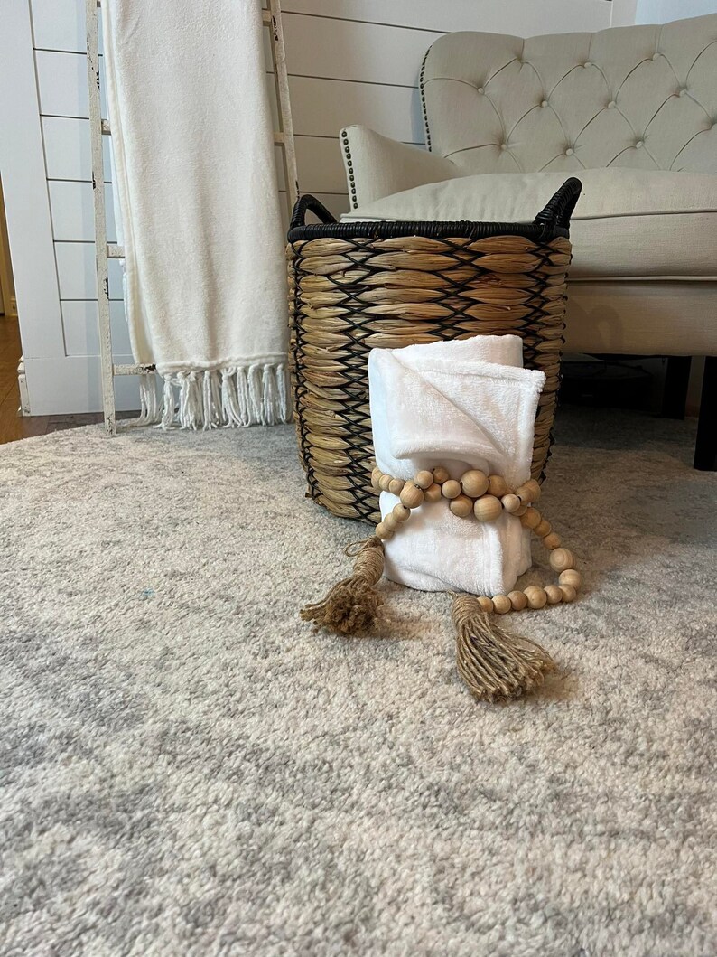 May include: A woven basket with a black rim and handles, filled with white towels and a wooden bead garland with tassels. The basket is sitting on a grey and white patterned rug.