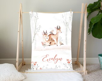 May include: A white baby blanket with a watercolor illustration of two deer and birds in a forest setting. The blanket has the name "Evelyn" written in cursive on the bottom.