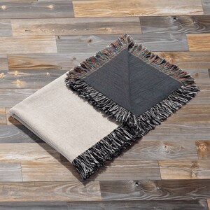 May include: A folded throw blanket with a white side and a dark grey side. The blanket has a fringe border and is lying on a wooden floor.
