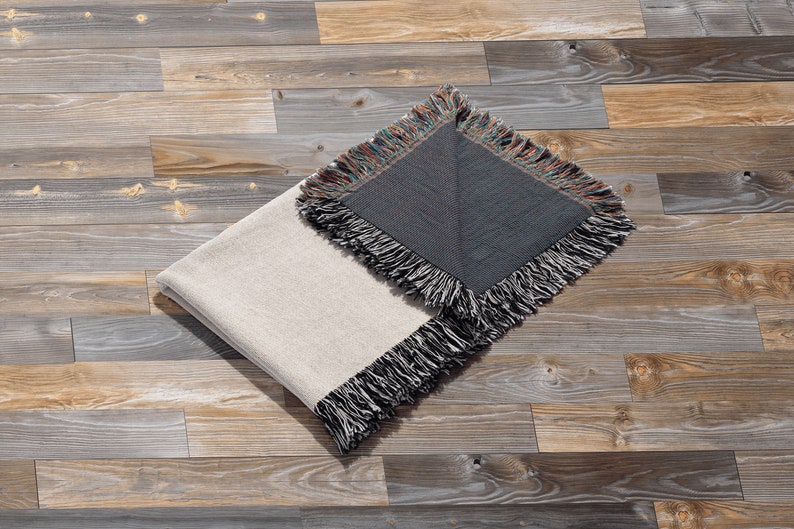 May include: A folded throw blanket with a white side and a dark grey side. The blanket has a fringe border and is lying on a wooden floor.