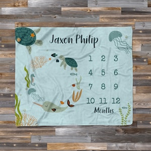 May include: A blue baby milestone blanket with a seahorse, narwhal, jellyfish, and sea turtles. The blanket has a number chart for each month of the year and the text "Jaxon Philip" at the top.
