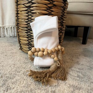 May include: A white, folded, soft fabric with a wooden bead garland and a brown tassel. The fabric is sitting on a light grey rug in front of a wicker basket.
