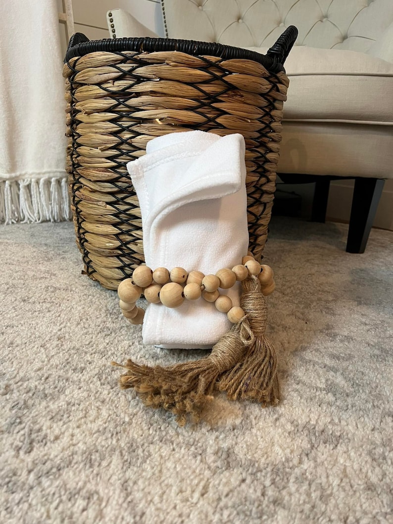 May include: A white, folded, soft fabric with a wooden bead garland and a brown tassel. The fabric is sitting on a light grey rug in front of a wicker basket.