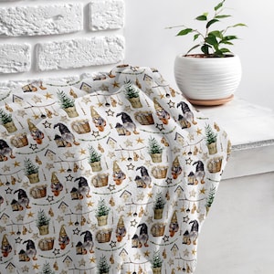 May include: White fabric with a repeating pattern of gnomes holding lanterns, stars, and small Christmas trees. The gnomes are wearing black hats and have white beards. The fabric is folded and draped over a white surface.