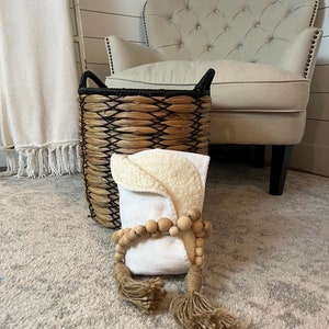 May include: A white throw blanket with tassels, a woven basket with a black rim, and a white cushion with a wooden bead garland and tassels. The items are on a light grey rug.