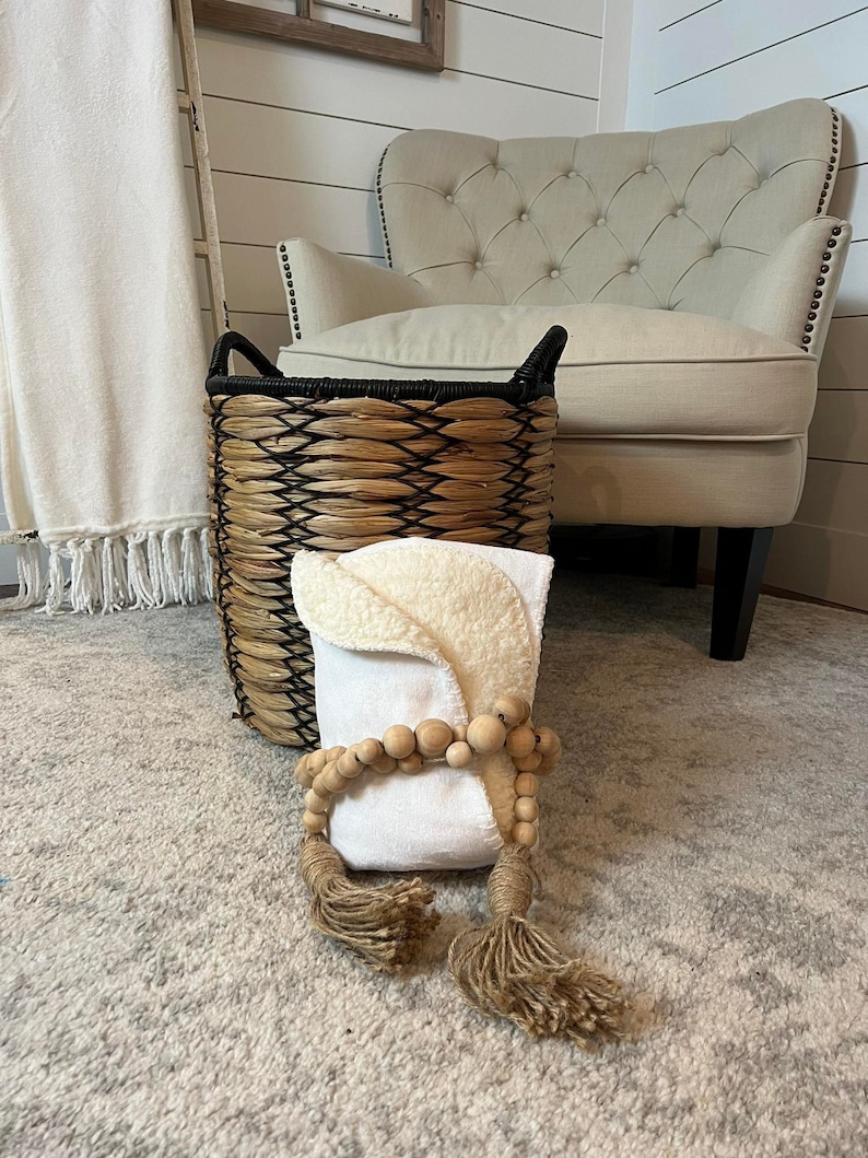 May include: A white throw blanket with tassels, a woven basket with a black rim, and a white cushion with a wooden bead garland and tassels. The items are on a light grey rug.