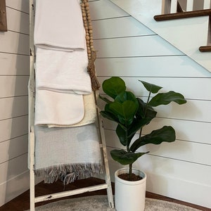 May include: A white wooden ladder leaning against a white wall with a white, grey, and brown blanket draped over it. A potted green plant sits on the floor next to the ladder.