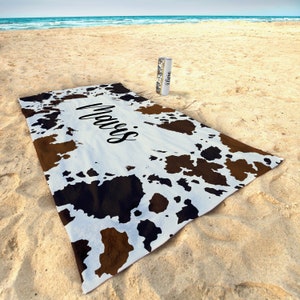 May include: A white beach towel with a brown and black cowhide pattern. The towel has the name "Mavis" written in black cursive lettering.