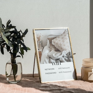 May include: A gold picture frame with a white mat and a photo of a bedroom interior. The frame is on a table with a vase of green leaves and a candle jar. The frame has the words "WIFI", "NETWORK", "Wifi Network", "PASSWORD", and "Wifi Password" printed on it.