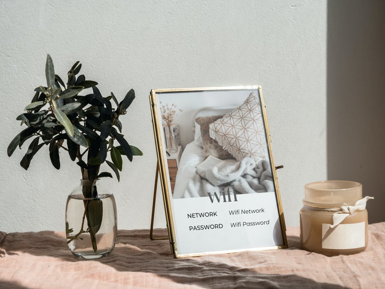 May include: A gold picture frame with a white mat and a photo of a bedroom interior. The frame is on a table with a vase of green leaves and a candle jar. The frame has the words "WIFI", "NETWORK", "Wifi Network", "PASSWORD", and "Wifi Password" printed on it.