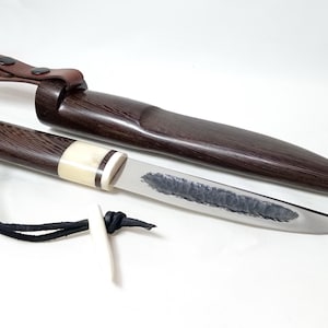 Könnte beinhalten: Ein feststehendes Messer mit einem Griff aus dunklem Holz, einer cremefarbenen Zwinge und einer polierten Stahlklinge mit texturierter, gehämmerter Oberfläche. Das Messer enthält eine passende Scheide aus dunklem Holz mit braunem Lederriemen.