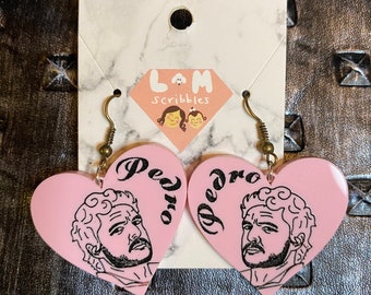 May include: A pair of pink heart-shaped earrings with a black and white illustration of Pedro Pascal on each earring. The earrings are labeled "Pedro" and are attached to gold-colored hooks.