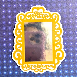 May include: A yellow framed sticker with a silver mirror inside. The frame has a decorative design and the text "the source of thy problems" is written on the bottom.